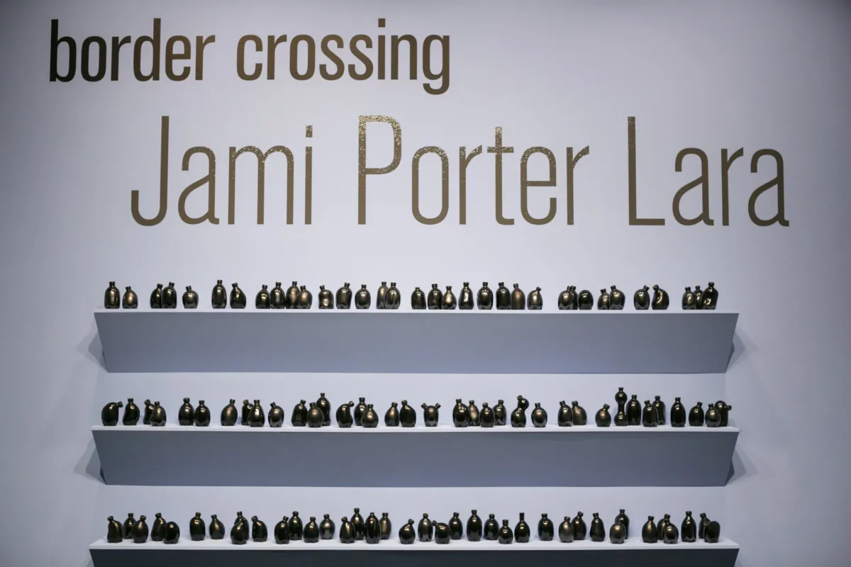 Shelves of small misshapen bottles in various shapes with the name of the exhibition above: Border Crossing: Jami Porter Lara above