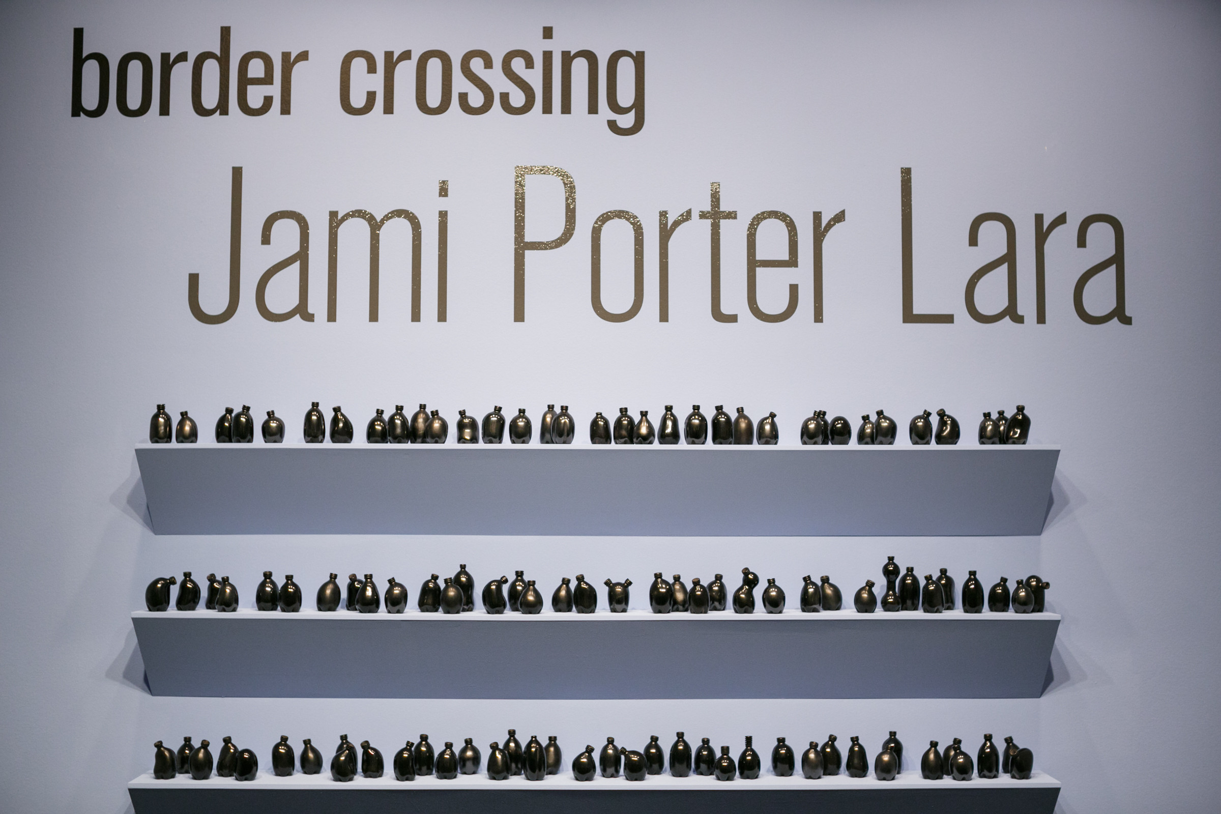 Shelves of small misshapen bottles in various shapes with the name of the exhibition above: Border Crossing: Jami Porter Lara above