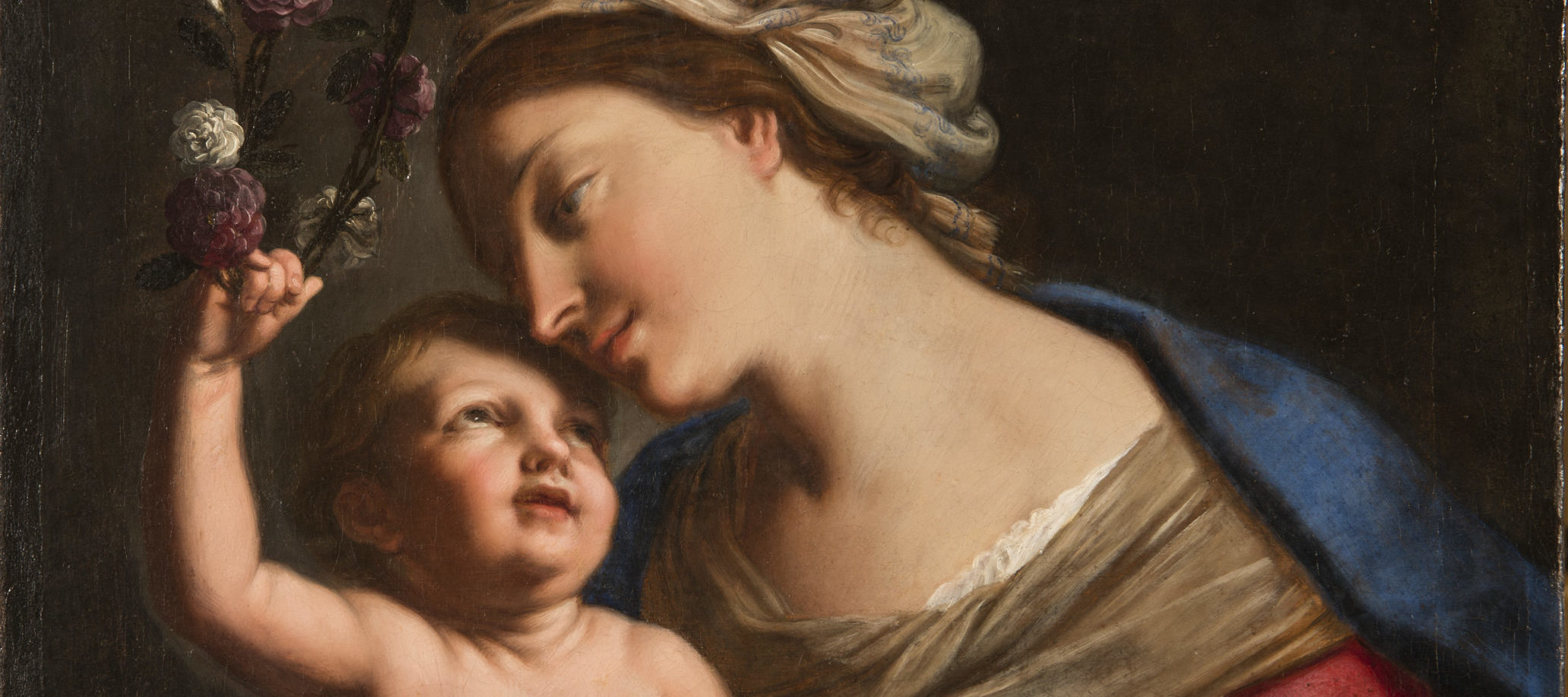 A young, light-skinned, brunette woman gazes down lovingly at the plump baby she holds on her lap. She wears a loose, tan turban, vivid blue cloak, and red dress with white sleeves. The light-skinned child returns her gaze, leaning back to crown the woman with a circlet of roses.