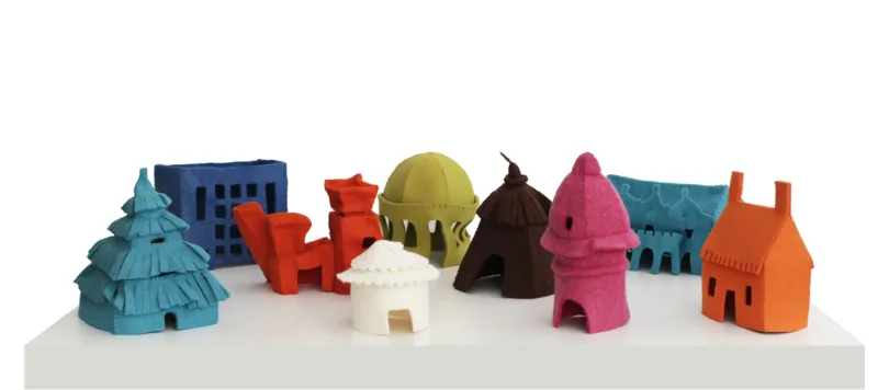 Arrangement of nine small, felt houses on a white table. Each house is a different, vibrant color and represents a different style of architecture spanning time and cultures—including a hut, a yurt, a cottage, art deco, postmodern architecture, and more.