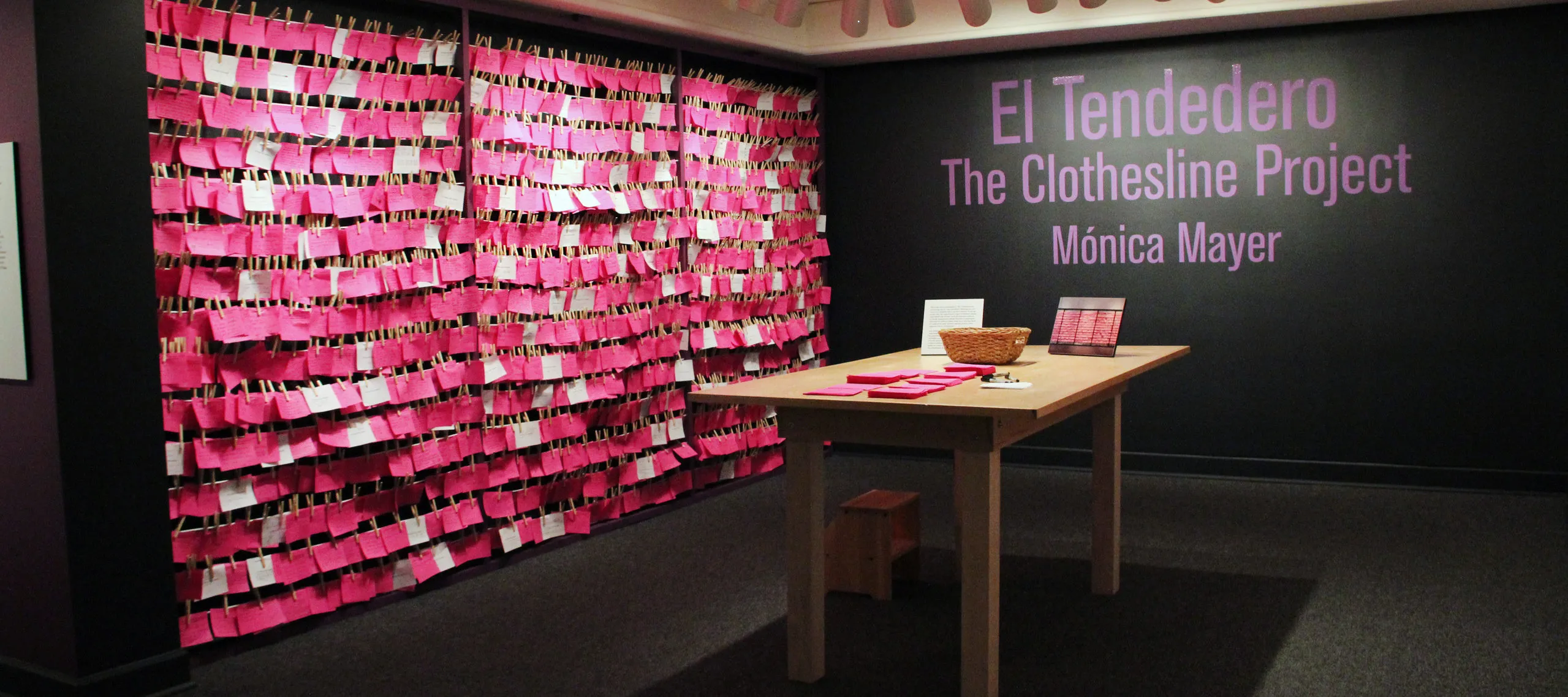 View of exhibition gallery features bright pink post-it notes line the wall from ceiling to floor. On the adjacent wall are the words in pink text:"El Tendedero, The Clothesline Project, Monica Meyer"