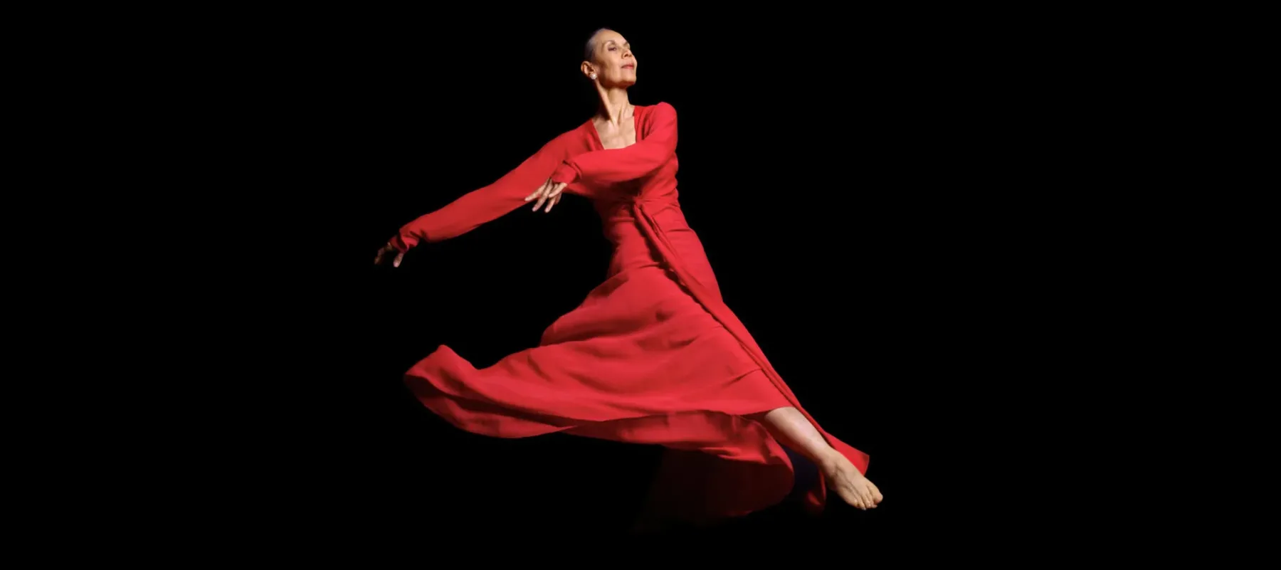 An older woman dances in a flowing red dress, appearing as if she's floating through space. Her face is turned to the right and she wears an expression of calm and fulfillment.