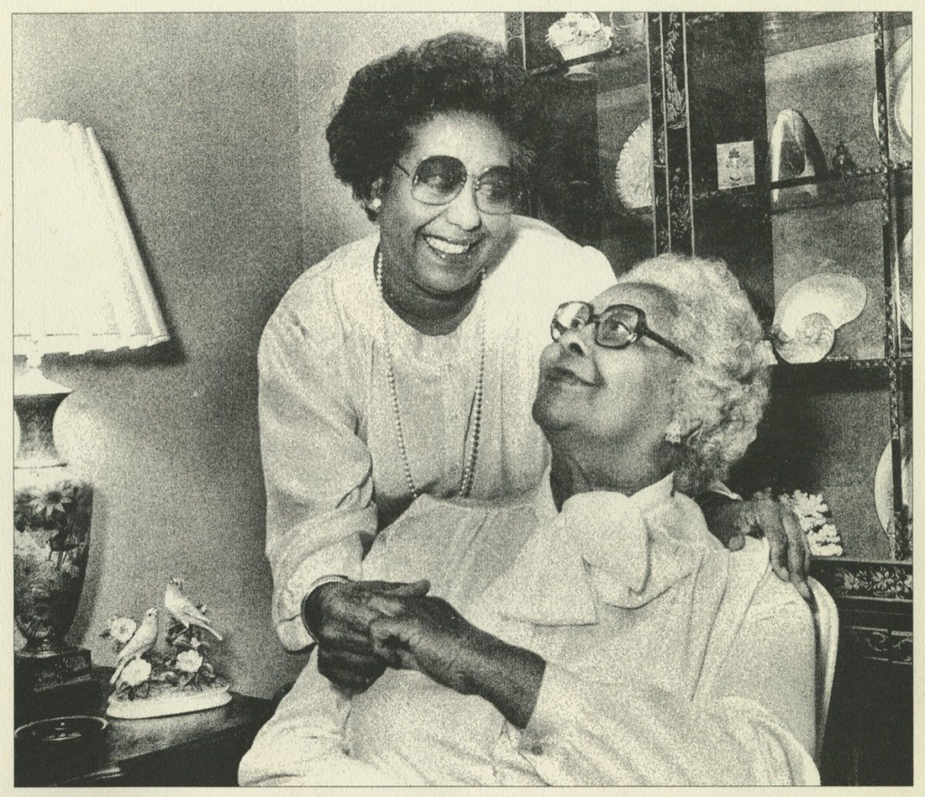 A black and white photograph in a home setting. One woman with large thick glasses and short white hair sits in a chair and looks up over her shoulder at the younger woman standing behind her. They hold hands; the young woman has her one arm around the older woman.