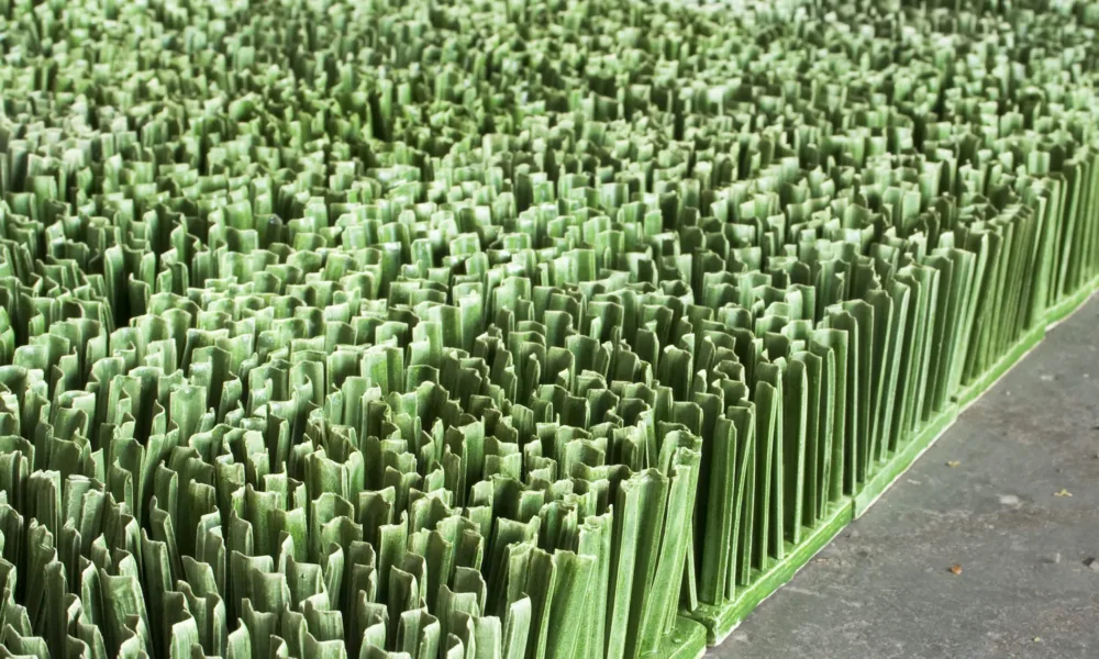 Detail photograph of ceramic sculpture made to look like a patch of lawn. Individual squares consisting of multiple upright blades of porcelain grass, glazed green, fit together to form a lush rectangular field of grass.