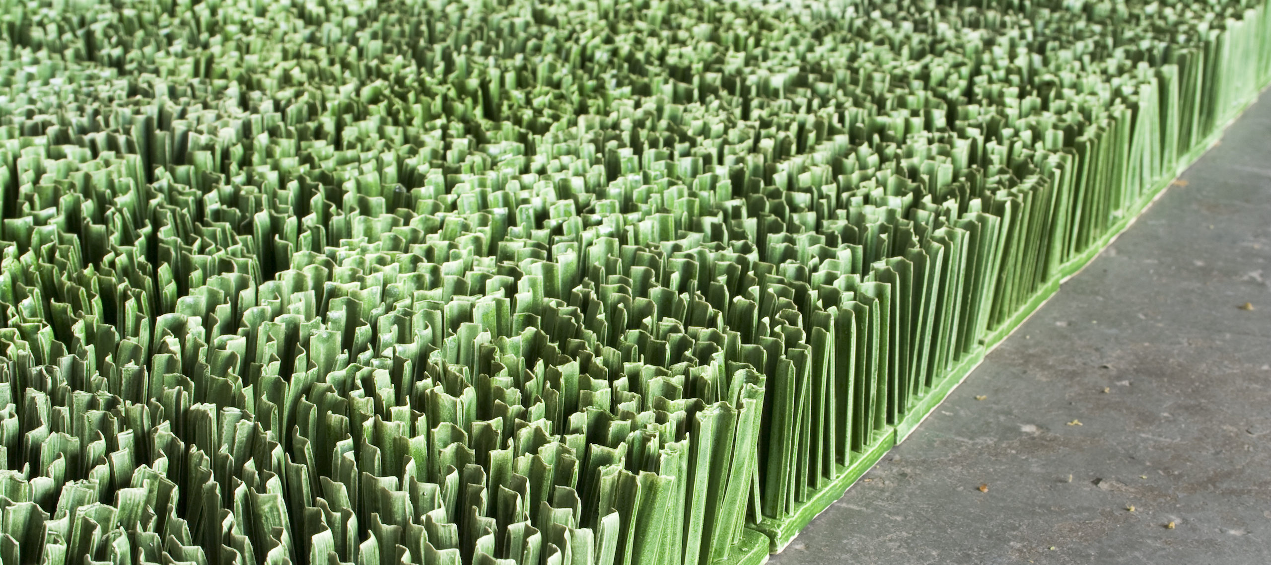 Detail photograph of ceramic sculpture made to look like a patch of lawn. Individual squares consisting of multiple upright blades of porcelain grass, glazed green, fit together to form a lush rectangular field of grass.