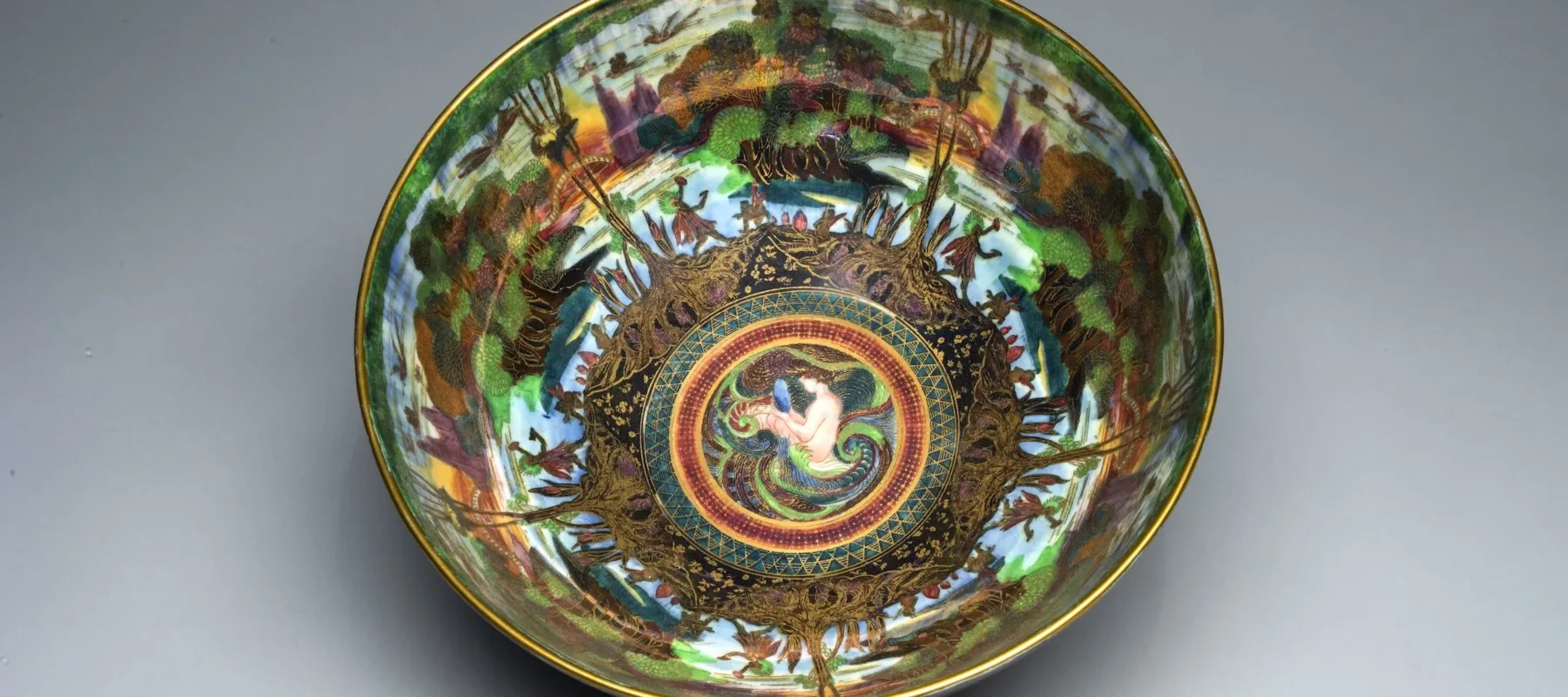 Interior view of a colorful, iridescent lusterware bowl featuring gold cloisonne and an ornamental Art Nouveau design. At the center, a light-skinned mermaid bathes in the ocean. Radiating outward, and repeated five times, is a lush agrarian scene.