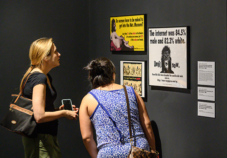 Two museum visitors are viewing art works hanging on a black wall. The art works are colorful and black-and-white posters with prints of gorillas and text.