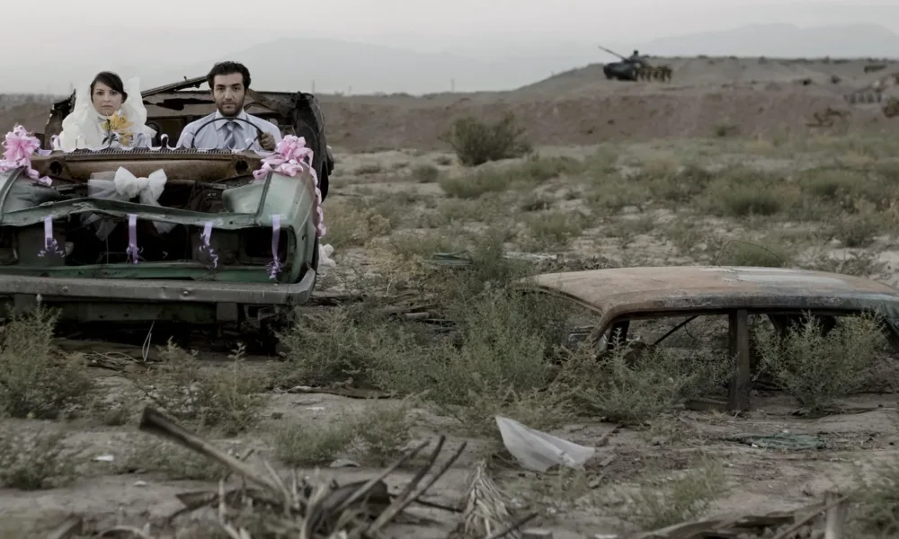 A couple sitting in a burned-out car in wedding finery, they look directly at the camera with neutral or stricken expressions.