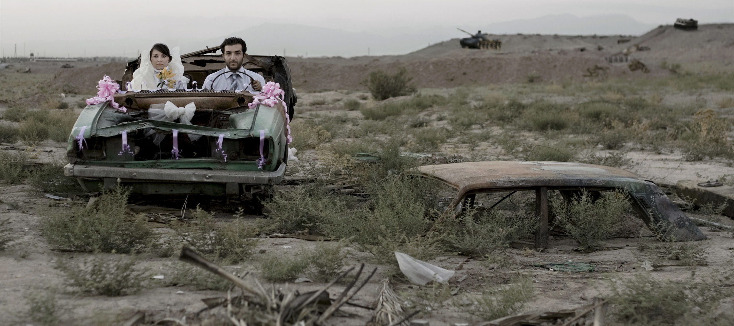 A couple sitting in a burned-out car in wedding finery, they look directly at the camera with neutral or stricken expressions.