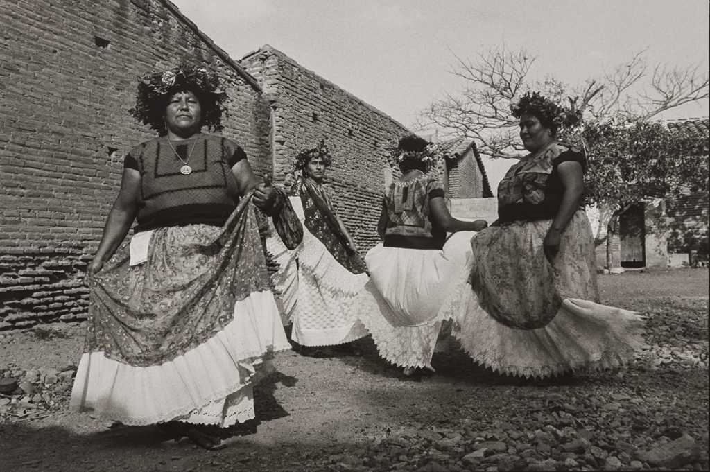 A black and white photograph of four medium-skinned women dancing. They wear floral headdresses and hold their long skirts, which are patterned with flowers and have large white hems, as they twirl in a rocky clearing next to a row of rural brick houses.