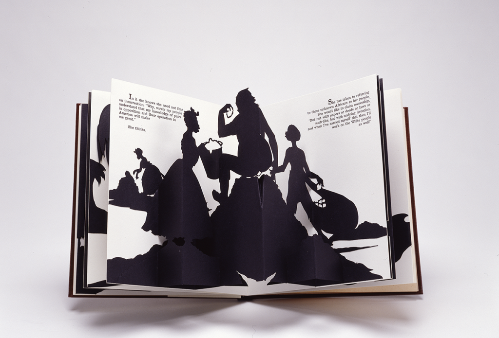 An open spread of a pop-up book featuring one of Walker's signature black-and-white cut-paper silhouettes which shows a female slave holding a bucket to, presumably, her master, who sits on, presumably, a big pile of cotton while a young boy slave is on the other side holding a bag of cotton. In the distance another female slave picks cotton in front of a heap. Text from the narrative is positioned on both the right and left pages in the upper corners.