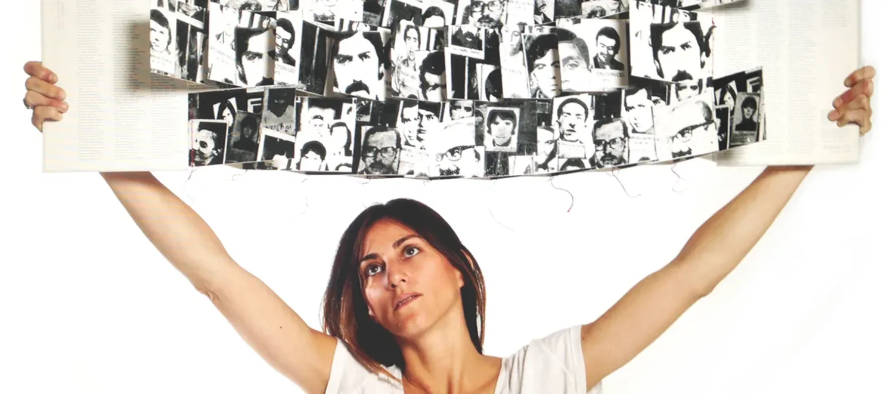 A woman with a light skin tone and brunette hair is holding up a white booklet with black-and-white photogrpaphs of a man with a mustache.