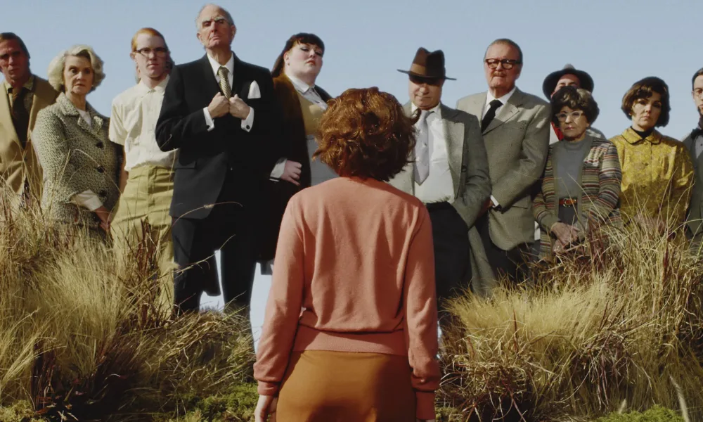 A color photograph of a woman with short red hair and pink cardigan, standing with her back to the camera in a field of tall yellow grass. She faces twelve men and women, who stand slightly elevated above her in a line and look pointedly at her.