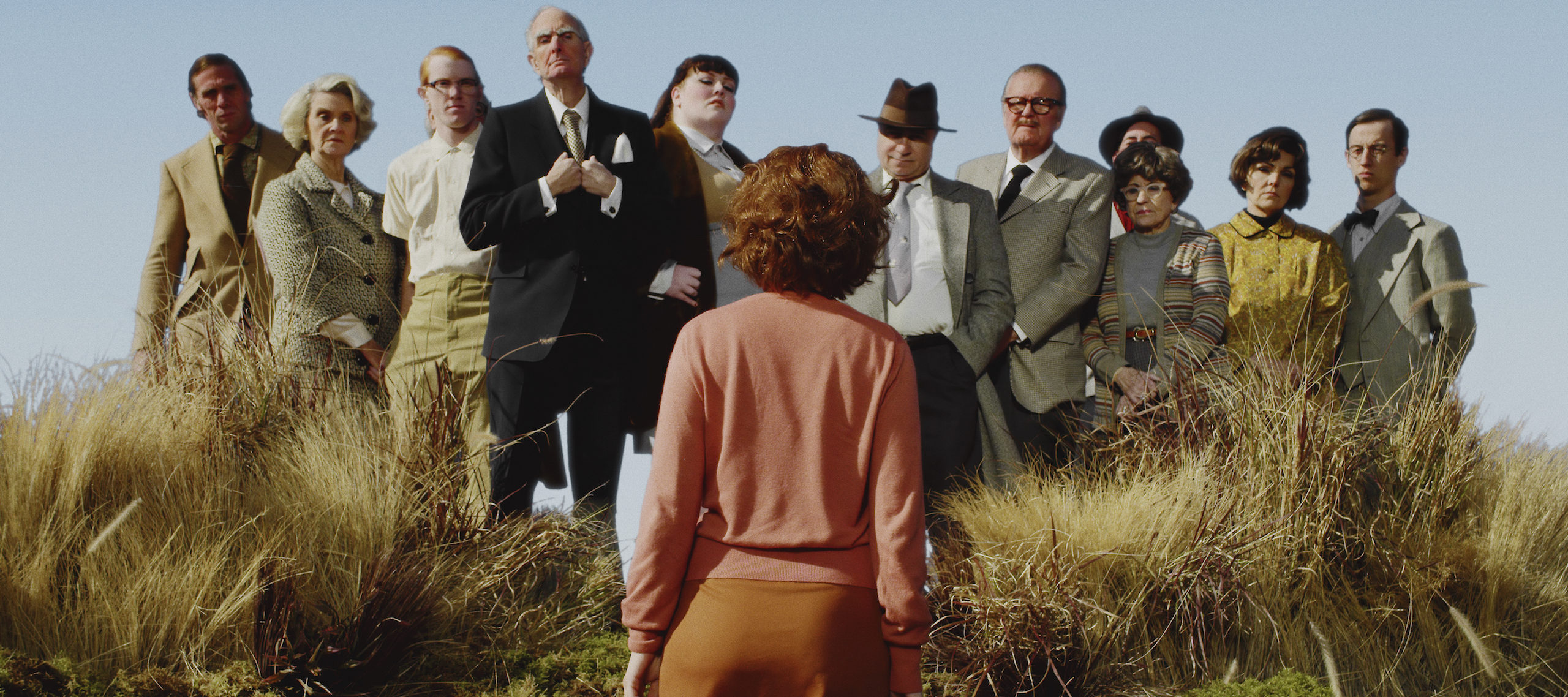 A color photograph of a woman with short red hair and pink cardigan, standing with her back to the camera in a field of tall yellow grass. She faces twelve men and women, who stand slightly elevated above her in a line and look pointedly at her.
