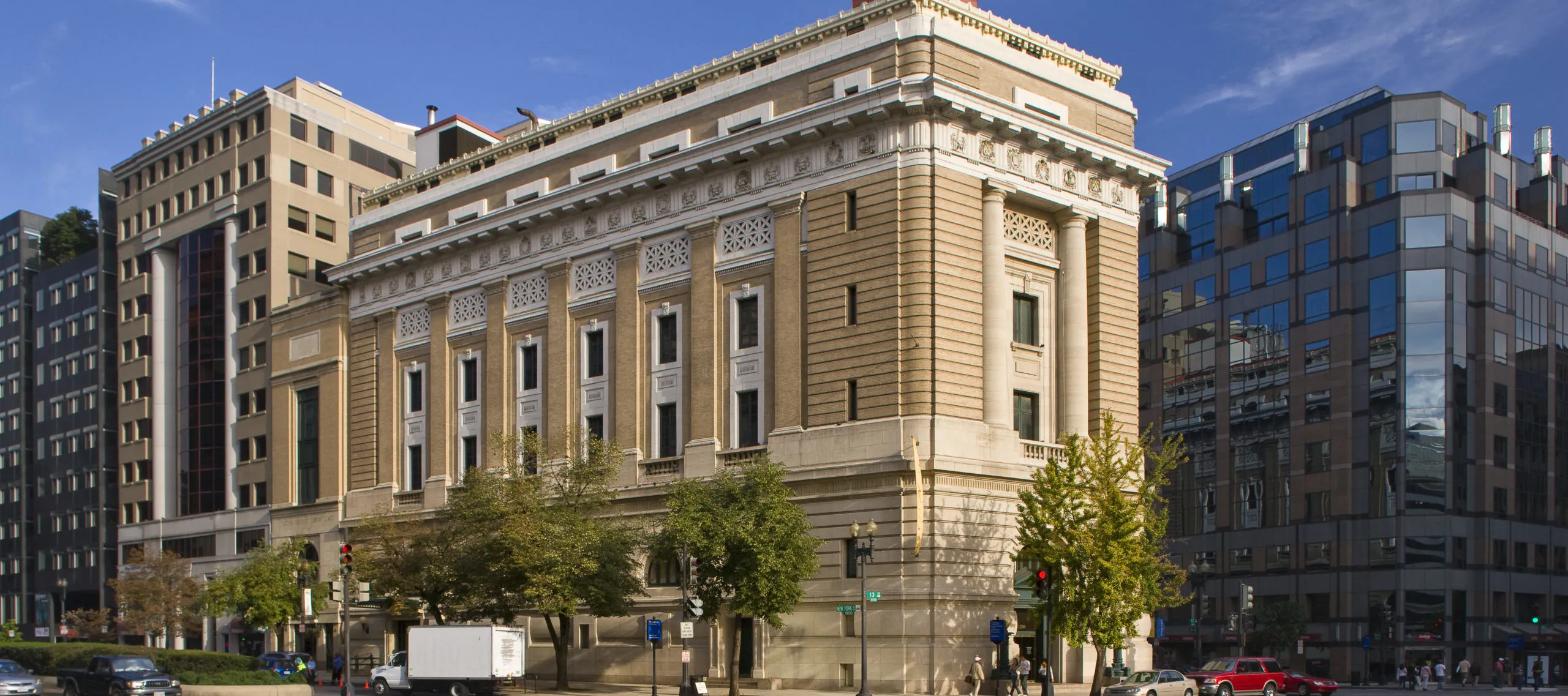 View of the museum from outside showing the Neoclassical building from one corner. The building is a tan-colored stone with an arched doorway, long vertical windows, and detailed molding around the roof.