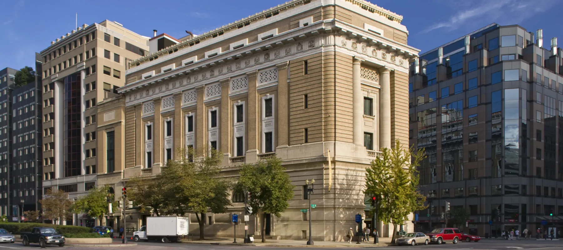 View of the museum from outside showing the Neoclassical building from one corner. The building is a tan-colored stone with an arched doorway, long vertical windows, and detailed molding around the roof.