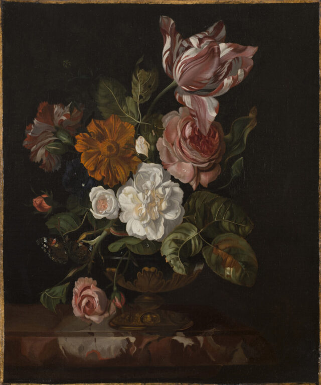 Pink, yellow, and white flowers in a golden vase are placed on a mahogany table against a muted, dark green background.