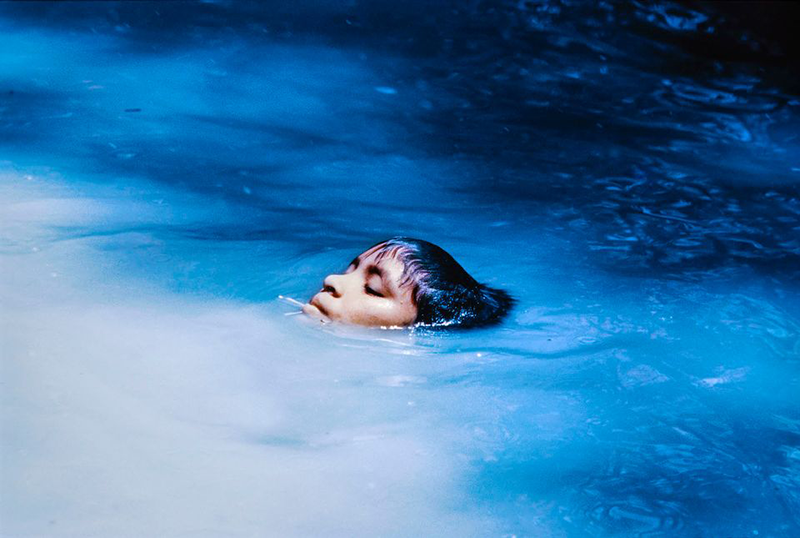 A color photograph of a young, medium-dark skinned person floating in water. The face of the youth, is in the center of the frame. The rest of their body is submerged under opaque, white-blue water that changes color in a gradient to dark blue in the upper right corner.