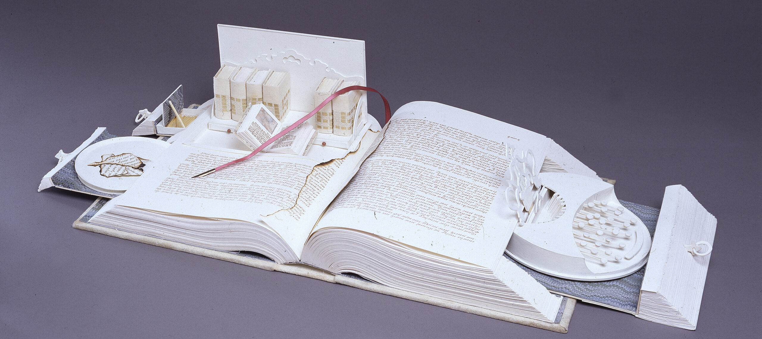 A white book opens like a Swiss army knife; its pages cut to create a sculpture. Opened to the center, pull-out drawers open from the sides, filled with paper-made objects. On the right, a typewriter-like object. On the left, a box with smaller books, including one that is also open.