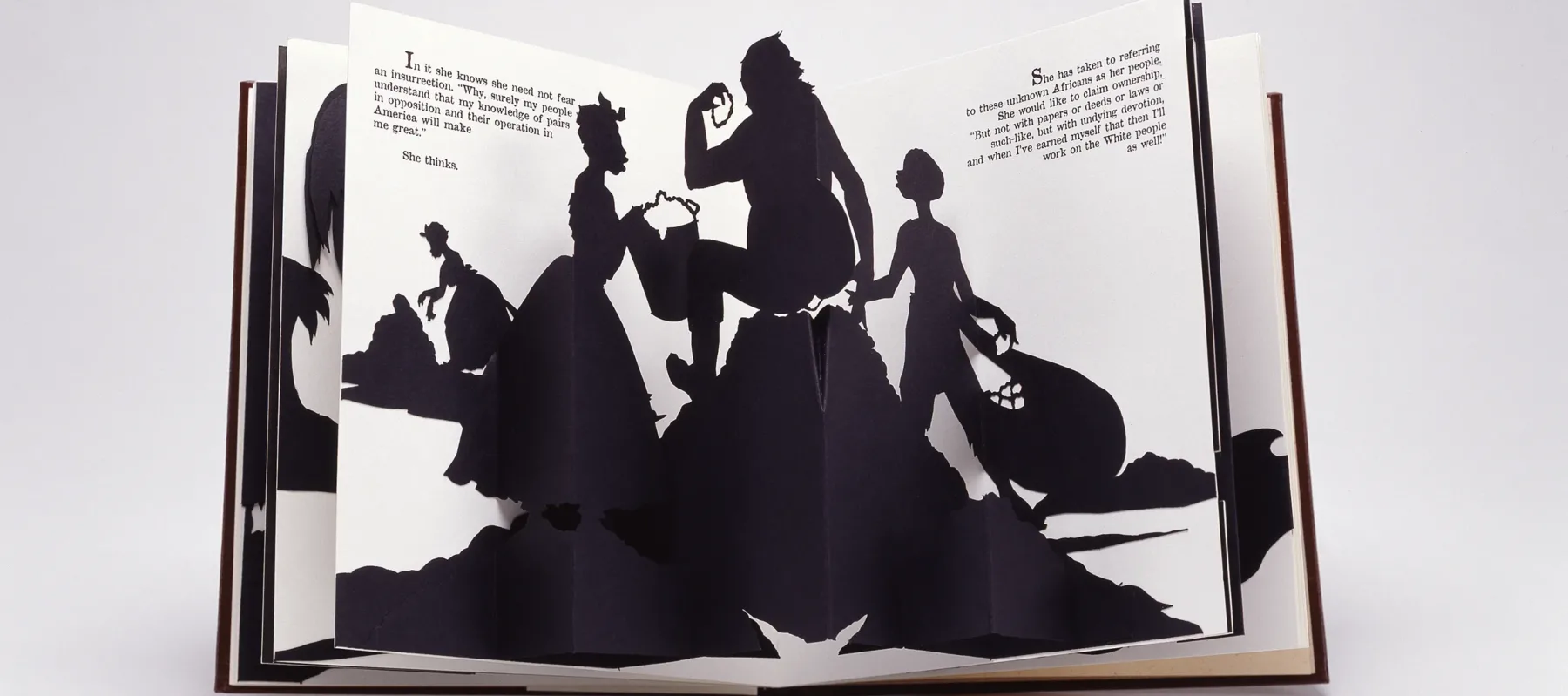 An open spread of a pop-up book featuring one of Walker's signature black-and-white cut-paper silhouettes which shows a female slave holding a bucket to, presumably, her master, who sits on, presumably, a big pile of cotton while a young boy slave is on the other side holding a bag of cotton. In the distance another female slave picks cotton in front of a heap. Text from the narrative is positioned on both the right and left pages in the upper corners.