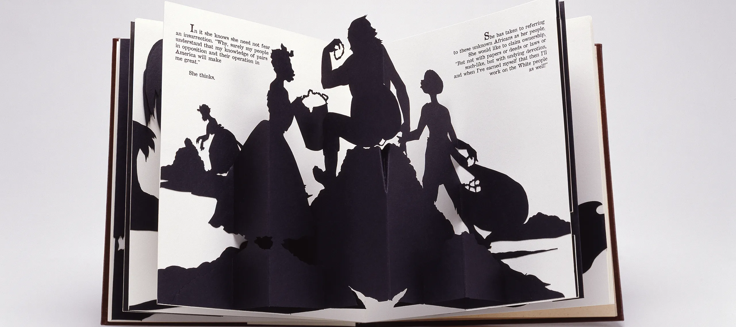 An open spread of a pop-up book featuring one of Walker's signature black-and-white cut-paper silhouettes which shows a female slave holding a bucket to, presumably, her master, who sits on, presumably, a big pile of cotton while a young boy slave is on the other side holding a bag of cotton. In the distance another female slave picks cotton in front of a heap. Text from the narrative is positioned on both the right and left pages in the upper corners.