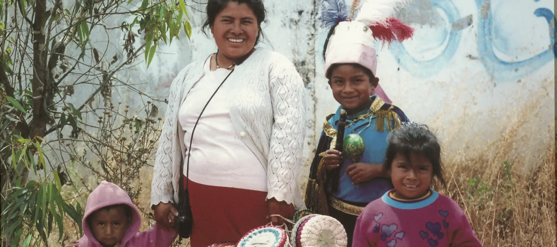 A medium-skinned woman smiles as she poses with three children among light brown shrubbery. She holds the hand of the smallest daughter and carries a bundle of colorful weaved baskets. A young boy wears a feathered white hat; the other young girl also carries baskets.