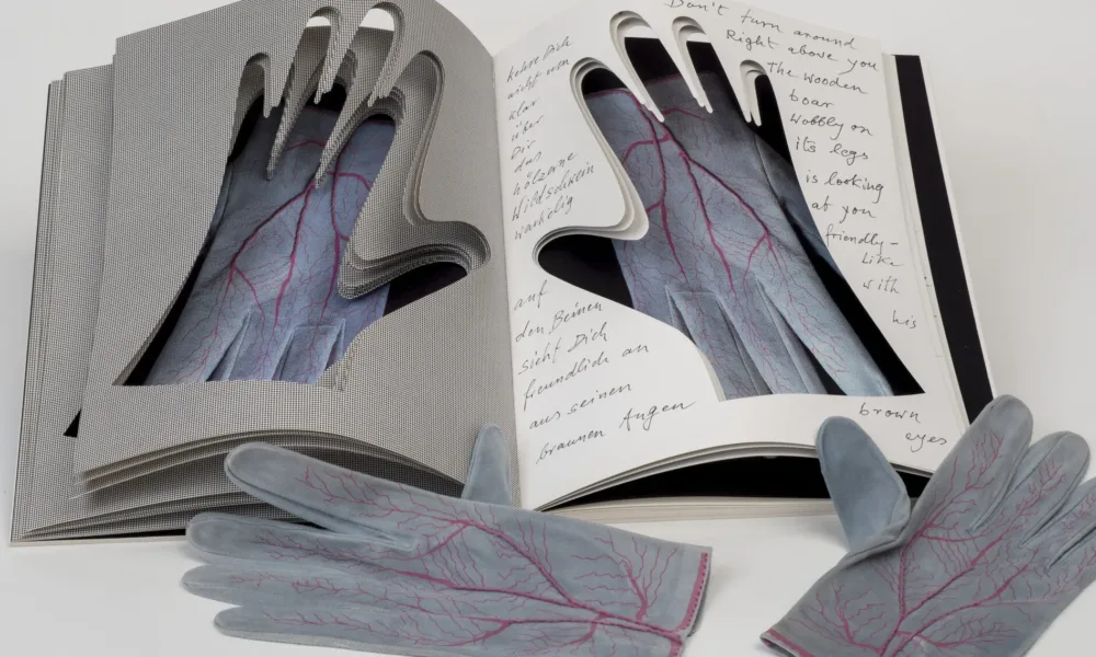 An open book into whose pages the shapes of hands have been cut. Gray gloves painted with red veins are visible through the cutouts. An identical pair of gloves rest next to the book.