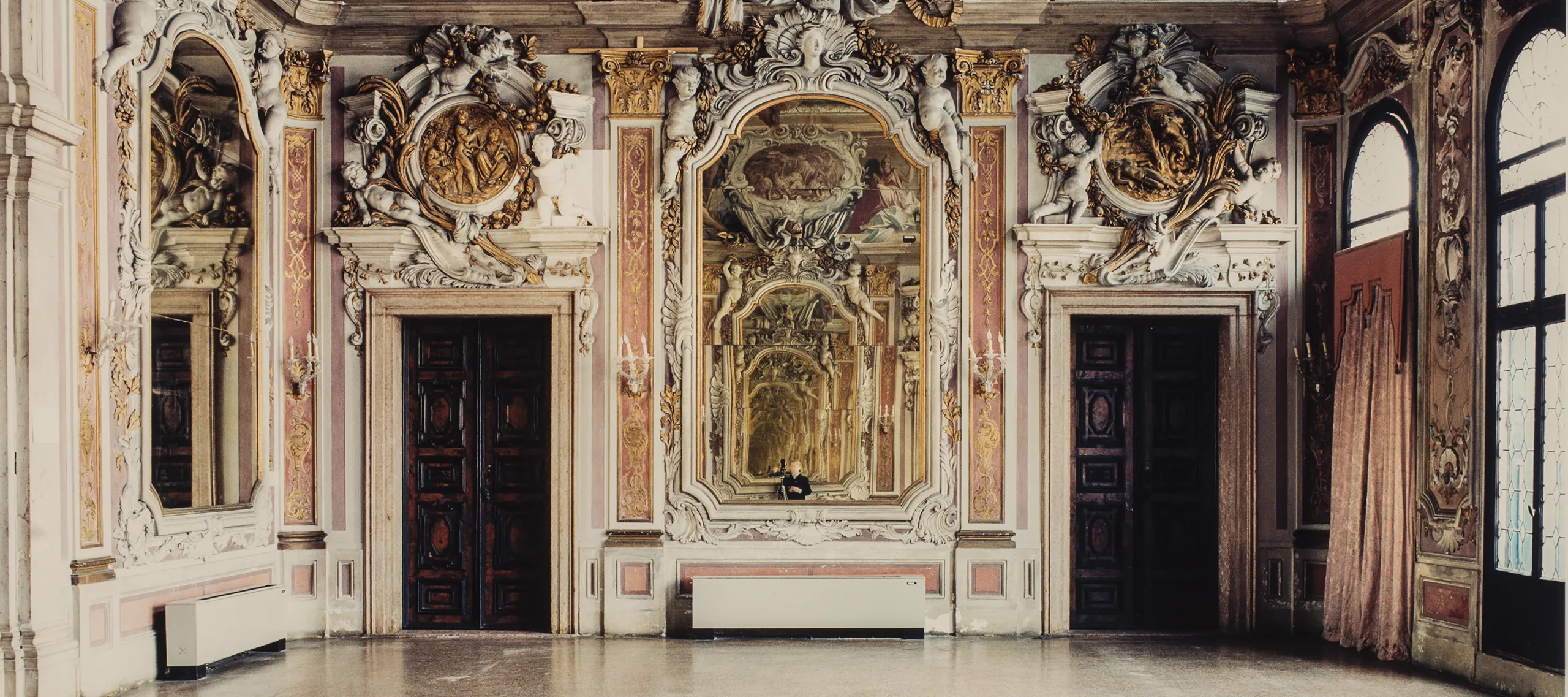 A color photograph of a large, ornate room in a palace. The expansive floor is bare and empty. At the other end of the room, a large ornate mirror hangs between two dark wooden doors. The moldings and details above the doors and mirror are extravagantly designed and a dramatic mural is featured at the very top of the wall. In the mirror, a small figure is seen, seemingly the photographer.