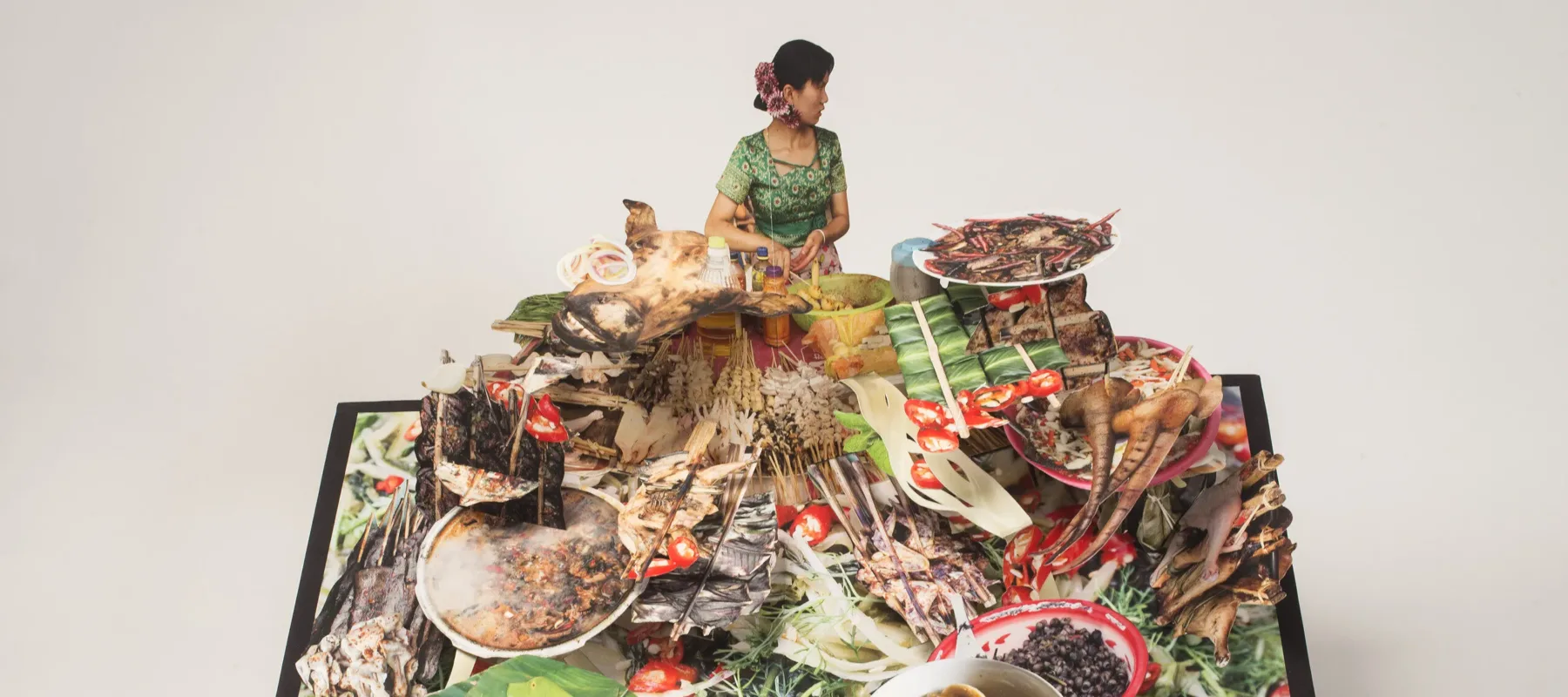 Large-scale pop-up book with a dark haired woman behind a huge display of food. She wears a green dress and purple flowers in her hair and turns to look over her left shoulder. The food explodes forward with bowls of soup, skewered meat, banana leaves, pig tail, and more.