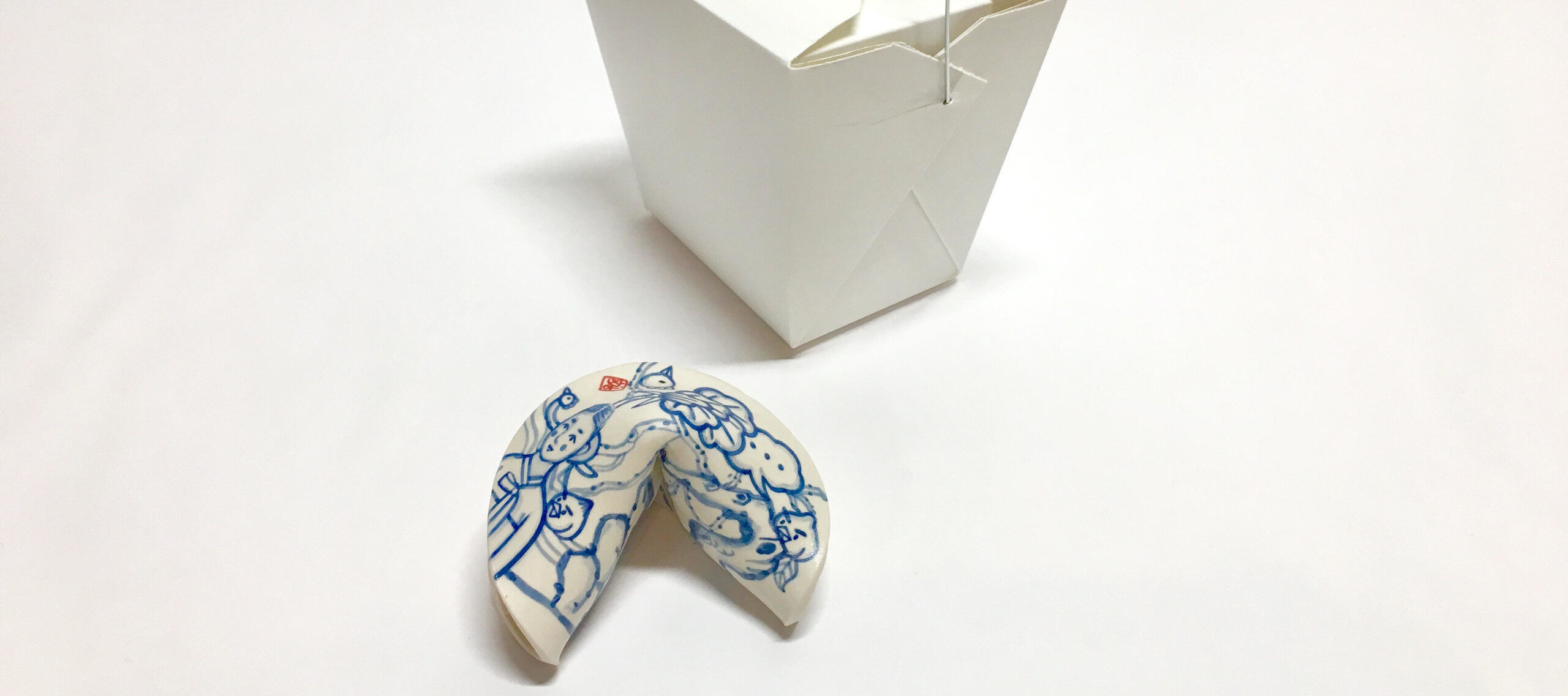 A small blue and white painted porcelain fortune cookie next to a white takeout box
