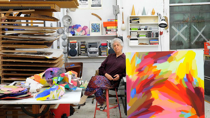 An elderly woman sits with her legs crossed on a stool. A large, colorful abstract canvas leans on a table in the foreground. Colorful pieces of paper are crumpled on a table next to it. Shelves on the walls are piled with canvases, sound systems, CD’s, tapes, books, and lamps.