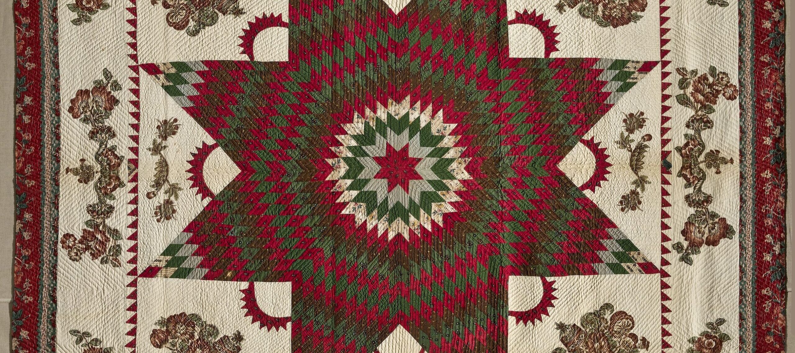 Vintage quilt in dark reds and greens on a cream background. In the center is a large eight-point Star of Bethlehem surrounded by Victorian floral appliques. The border, edged in dark red fabric, features matching floral garland appliques.