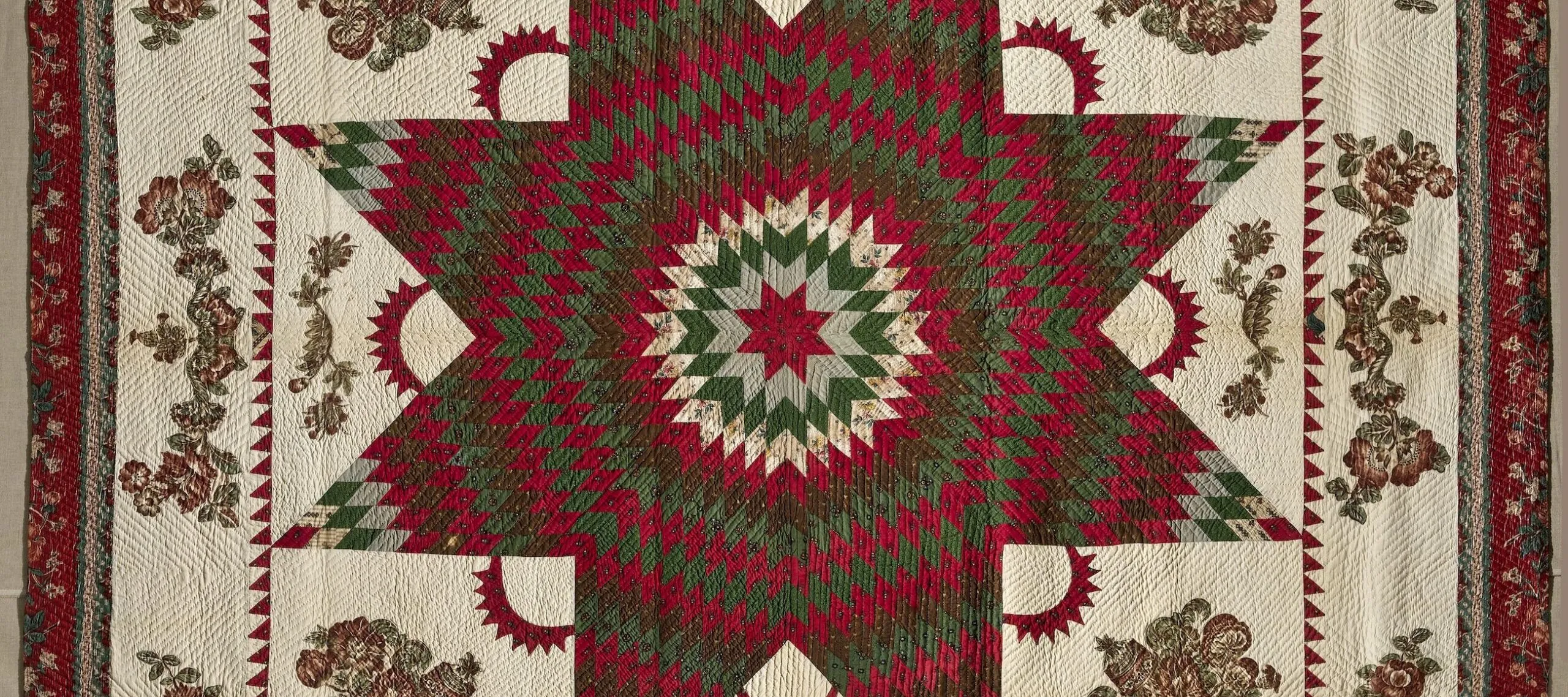 Vintage quilt in dark reds and greens on a cream background. In the center is a large eight-point Star of Bethlehem surrounded by Victorian floral appliques. The border, edged in dark red fabric, features matching floral garland appliques.