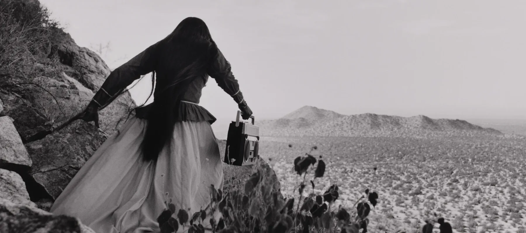 A black-and-white photograph shows the back of a woman as she crests a rocky path above a vast desert landscape beneath an expansive sky. Her traditional, ethnic full skirt, long-sleeved blouse, and long, straight, dark hair contrasts with the modern portable stereo she carries.