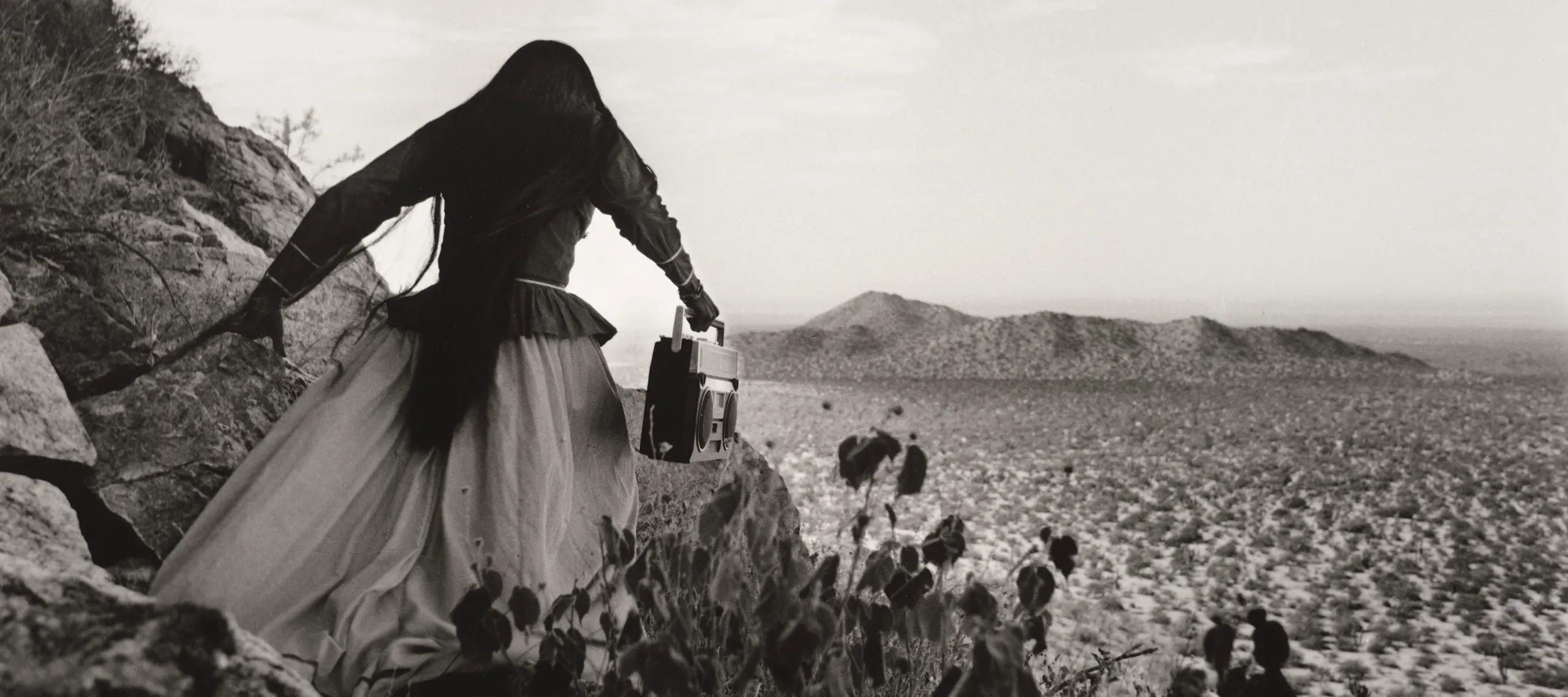 A black-and-white photograph shows the back of a woman as she crests a rocky path above a vast desert landscape beneath an expansive sky. Her traditional, ethnic full skirt, long-sleeved blouse, and long, straight, dark hair contrasts with the modern portable stereo she carries.