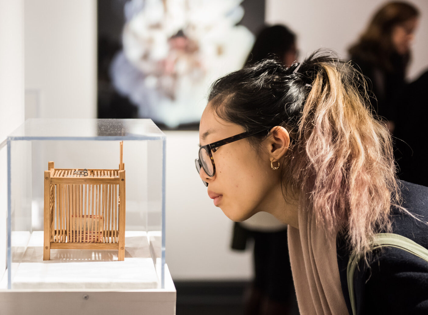 Partial gallery view of a young woman in a ponytail and glasses bending down to look at a tiny contemporary sculpture in a glass vitrine. The sculpture is a wooden cage with an object stored inside. On the back wall hangs a photograph of a light-skinned woman in a white dress.