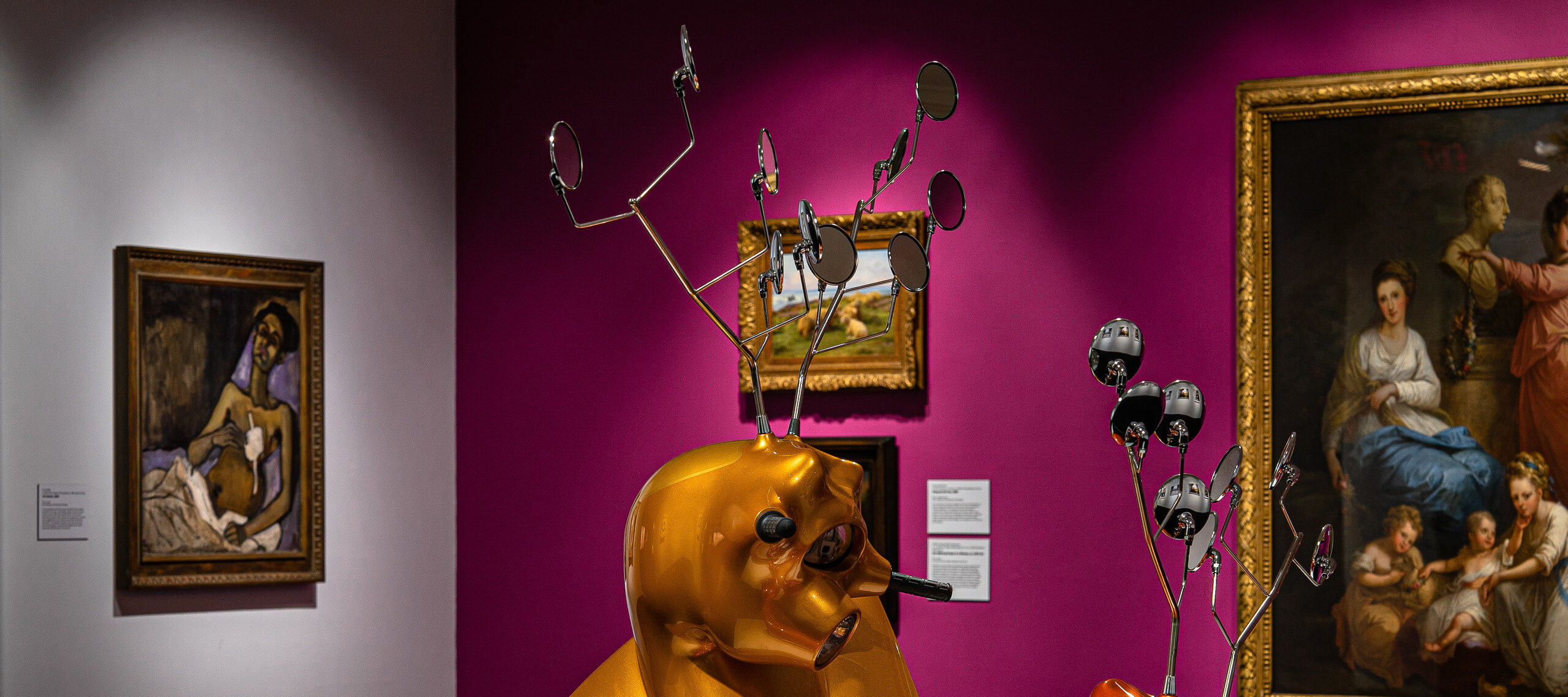 Gallery view with contemporary sculpture made of motorscooters that resemble two antlered animals fighting in foreground.Three paintings hang on a magenta wall in background. To the left on a white wall hangs another painting.