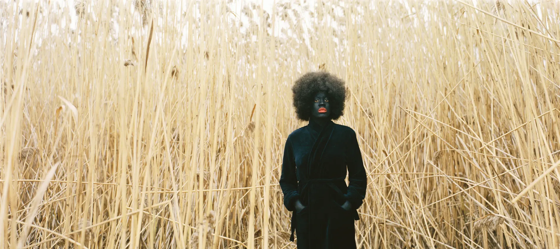 Xaviera Simmons (b. 1974) staged herself among a towering thicket of yellow reeds in her photograph One Day and Back Then (Standing) (2007). Simmons wears a black trench coat, black tights and boots, black face makeup, and bright red lipstick—a presentation that starkly juxtaposes her form against the landscape.