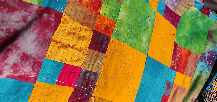 A close-up image of a color block quilt, taken diagonally. Colors repeat throughout without a set pattern. A golden yellow is the predominant color, but there are also lush light green, aqua, magenta, and tan blocks; some blocks look to be tie-dyed in multiple colors.