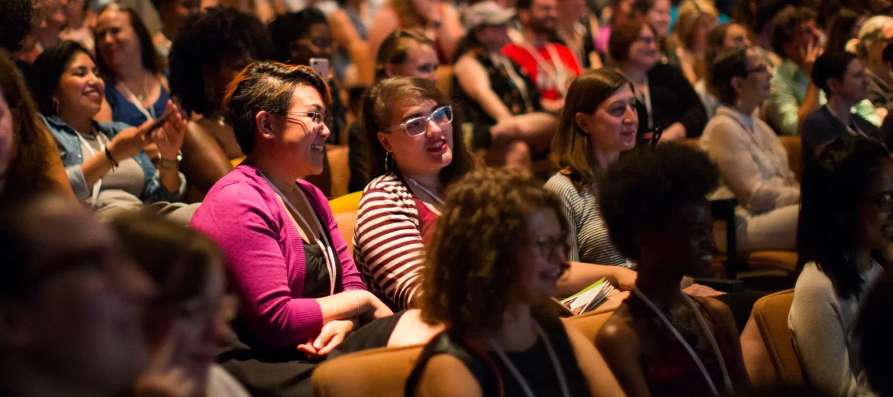 A color photograph of a brightly lit auditorium. Smiling women take up almost every seat. Some are taking photos of the stage with their phone; others lean together in conversation.