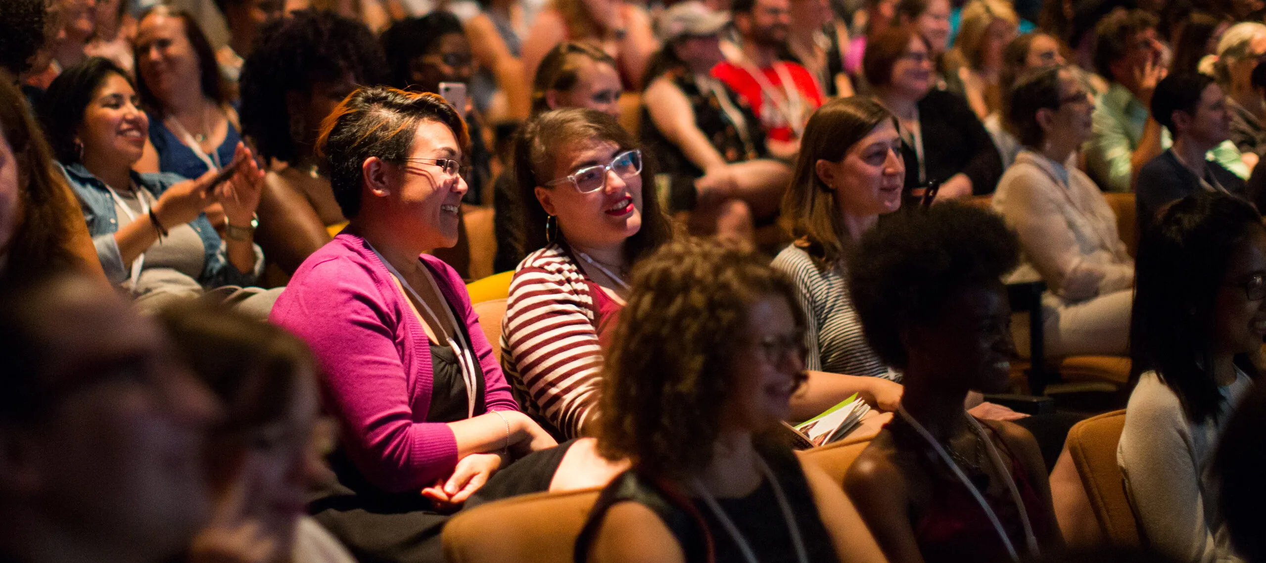 A color photograph of a brightly lit auditorium. Smiling women take up almost every seat. Some are taking photos of the stage with their phone; others lean together in conversation.