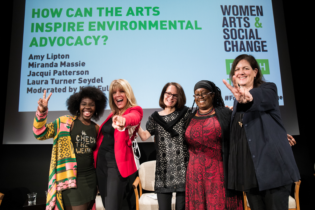 Five diverse women pose happily in NMWA's performance hall, in front of a projected slide that says "How can the arts inspire environmental advocacy?" They smile wide, one woman points happily at the camera, and another throws up a peace sign. 