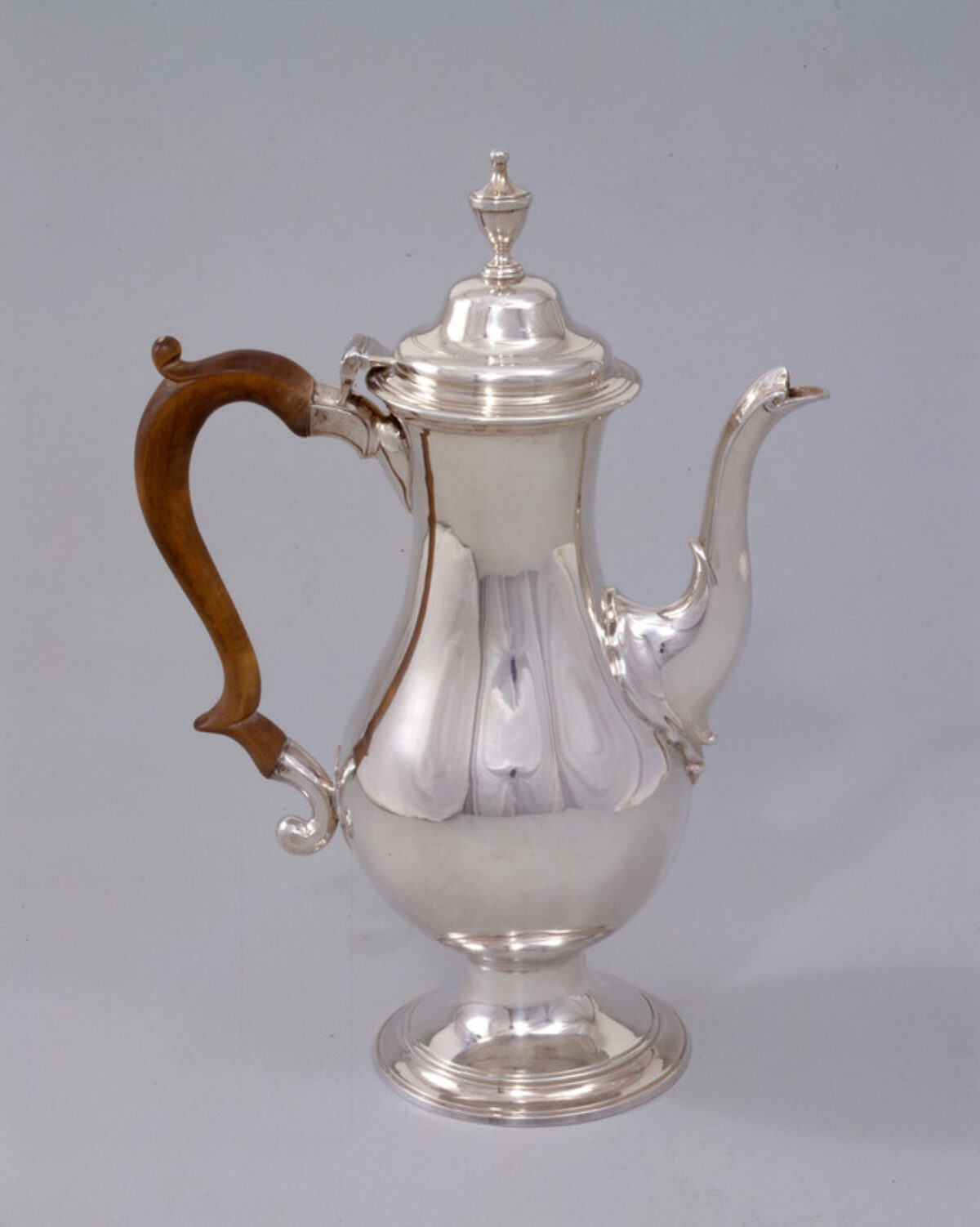Silver coffee pot features a tall, baluster-shaped body, swan-neck spout, and a wooden scroll handle. In keeping with the Neoclassical style, the pot is smooth, simple and relatively unadorned.