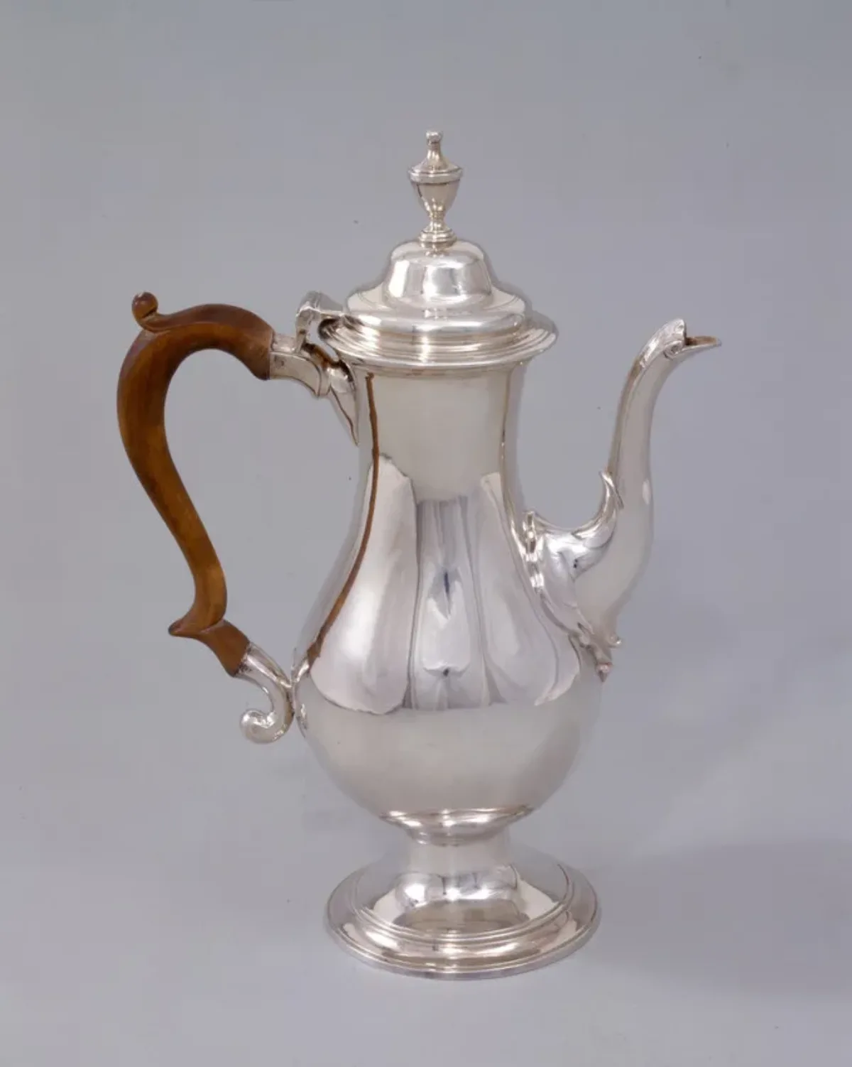 Silver coffee pot features a tall, baluster-shaped body, swan-neck spout, and a wooden scroll handle. In keeping with the Neoclassical style, the pot is smooth, simple and relatively unadorned.