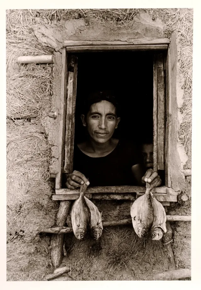 A black-and-white photograph features an open, rough-hewn window framing the head and shoulders of a woman with dark hair and medium-dark skin. Peering out from a murky interior, she displays 2 fish per hand on the window ledge. A mud-grass mixture textures surrounding walls.