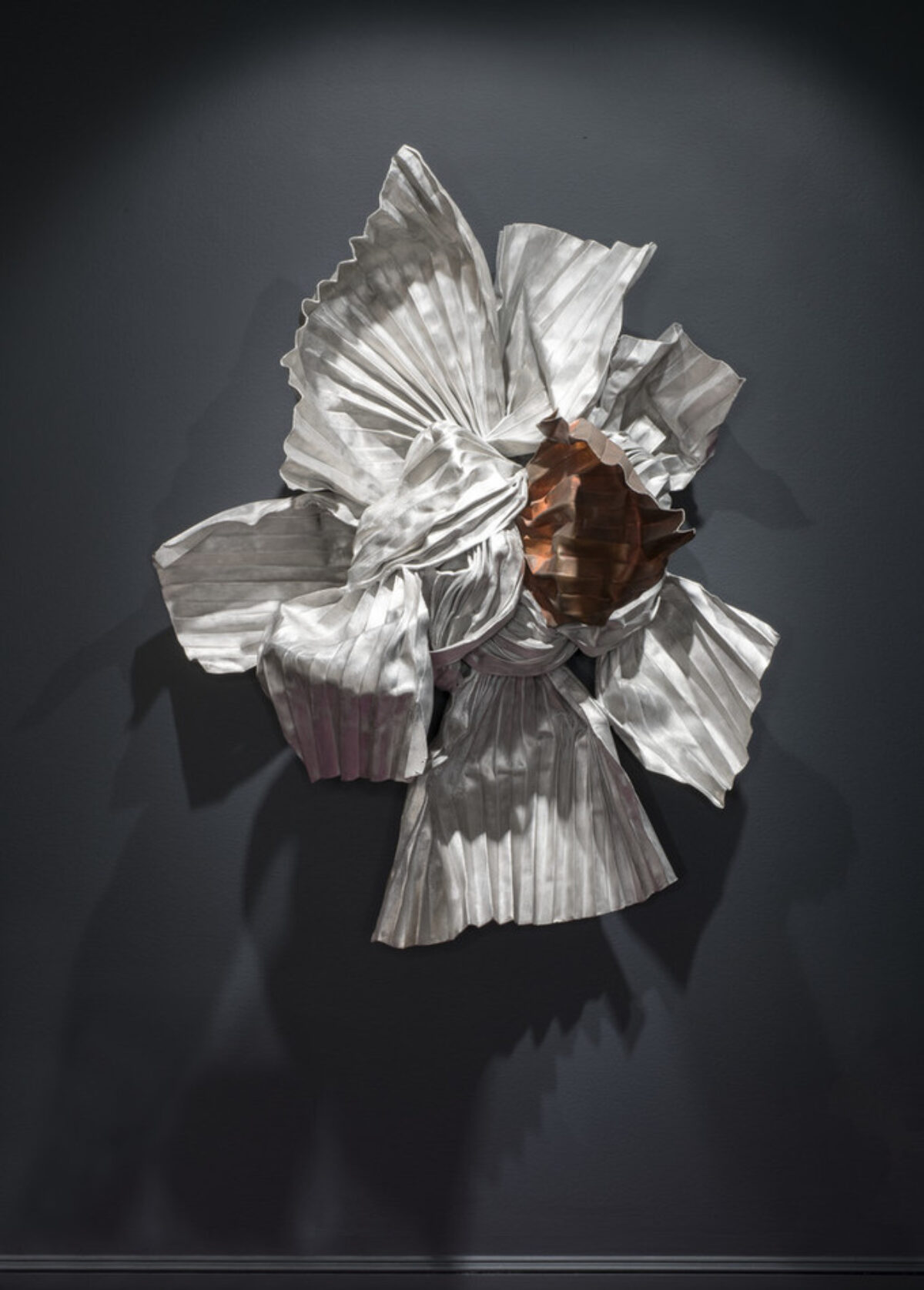 A wall-mounted sculpture made of metal convincingly resembles an elaborate knot of twisted, rolled, and tied silver and copper fabrics. Pleated ends radiate at different angles from the tangled, coppery center and project out from the wall into the viewer’s space.