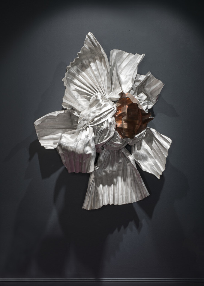 A wall-mounted sculpture made of metal convincingly resembles an elaborate knot of twisted, rolled, and tied silver and copper fabrics. Pleated ends radiate at different angles from the tangled, coppery center and project out from the wall into the viewer’s space.