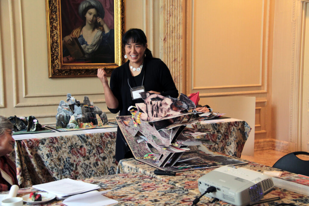 An Asian woman with a light skin tone, black hair tied back and white, round-shaped white jewellery is standing next to a large, open pop-up book, smiling.