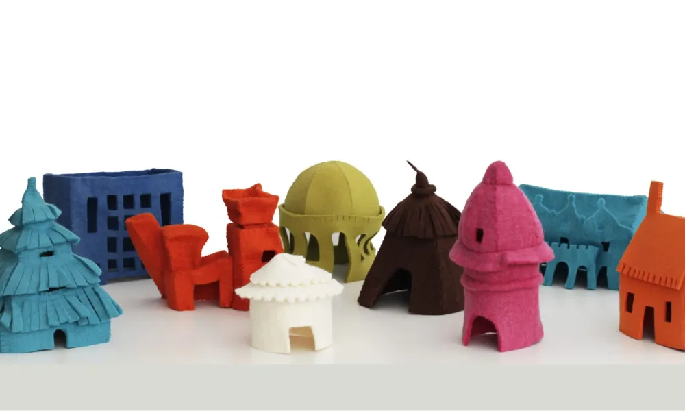 Arrangement of nine small, felt houses on a white table. Each house is a different, vibrant color and represents a different style of architecture spanning time and cultures—including a hut, a yurt, a cottage, art deco, postmodern architecture, and more.