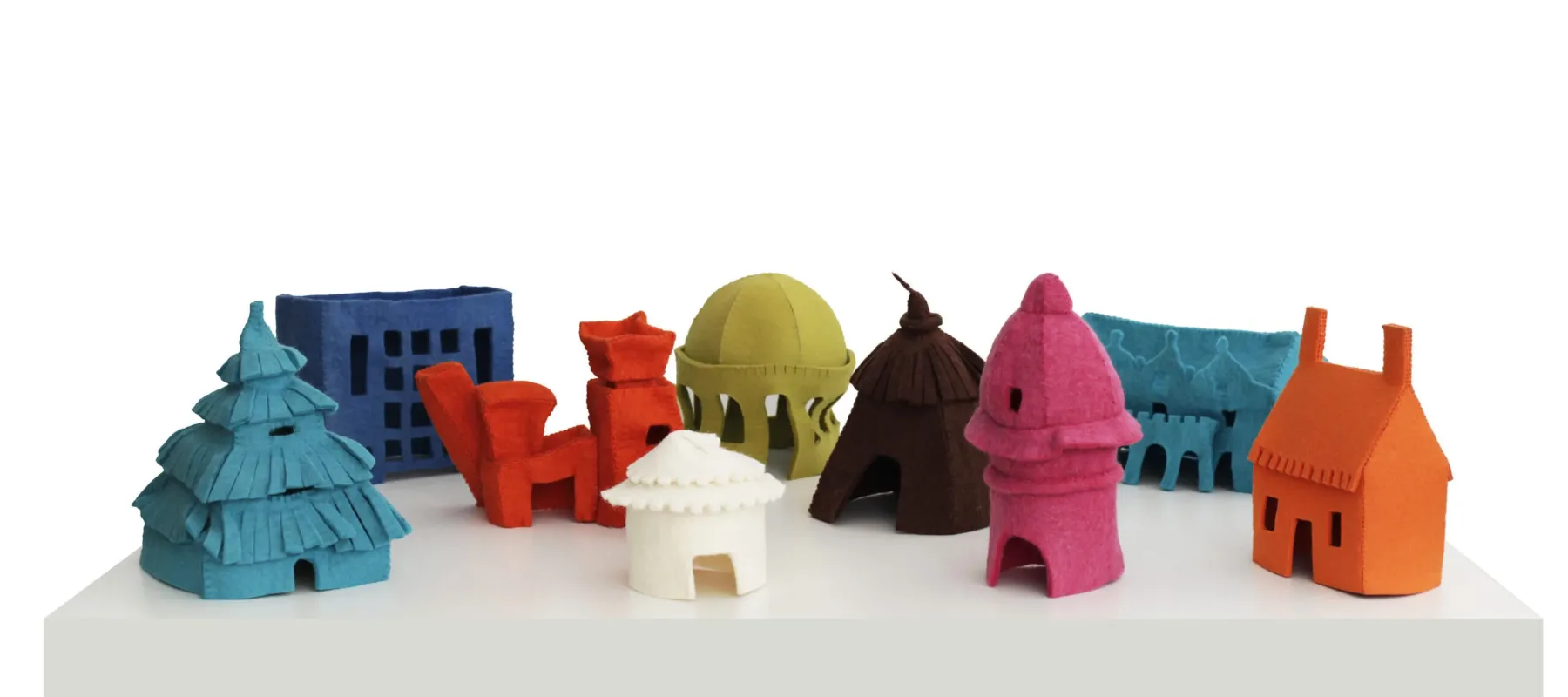 Arrangement of nine small, felt houses on a white table. Each house is a different, vibrant color and represents a different style of architecture spanning time and cultures—including a hut, a yurt, a cottage, art deco, postmodern architecture, and more.